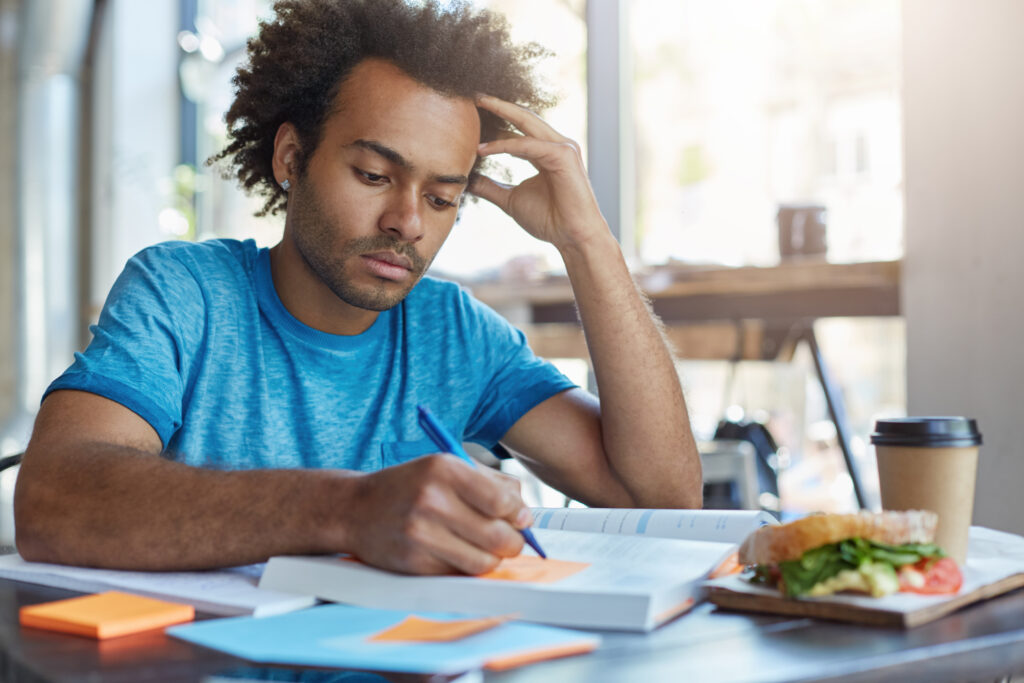 Handsome serious black European male student busy learning lessons during lunch at cafe, sitting at table with food and textbooks, making notes, writing down new words preparing for Spanish class